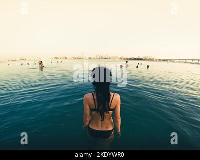 Rückansicht Junge kaukasische Frau tritt an heißen Tagen im UAE Aquaventure Park im Privatstrand des Atlantis Hotels in das Meerwasser ein, um dort zu schwimmen Stockfoto