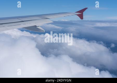 Der Blick vom Fenster des Flugzeugs auf den Flügel des Flugzeugs vor dem Hintergrund von flauschigen lockigen Wolken und blauem Himmel. Das Konzept des Reisens. Stockfoto