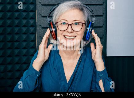 Behandlung von Hörverlust. Positive reife Frau mit Audiometrie-Kopfhörern beim Hörtest und Audiogramm in einem speziellen Audioraum Stockfoto