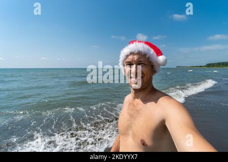 Lächelnder Mann in einem Weihnachtsmannhut nimmt ein Selfie vor dem Hintergrund des Meeres und kopiert den Raum. Weihnachtsferien in warmen Ländern Stockfoto