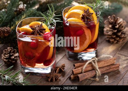 Glühwein mit Orangenscheiben, Preiselbeeren und Gewürzen auf rustikalem Holztisch mit Christbaumschmuck im Hintergrund Stockfoto