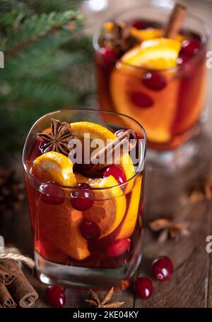 Glühwein mit Orangenscheiben, Preiselbeeren und Gewürzen auf rustikalem Holztisch mit Christbaumschmuck im Hintergrund Stockfoto