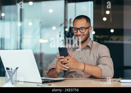 Glücklicher afroamerikanischer Geschäftsmann mit Brille, der im modernen Büro mit einem Laptop arbeitet, Mann im Hemd sitzt am Tisch und stöbert mit dem Smartphone auf Online-Seiten. Stockfoto