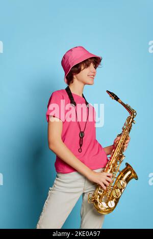 Portrait eines fröhlichen Teenagers mit lockigem, braunem, kurzen Haar, das in rosa panama posiert und auf blauem Hintergrund Saxophon spielt Stockfoto