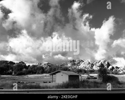 Eine atemberaubende Graustufenaufnahme der flauschigen Wolkenlandschaft über dem ländlichen Feld mit kleinen Gebäuden und felsigen Hügeln Stockfoto