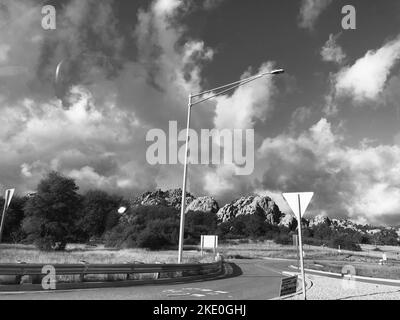Eine Graustufenaufnahme einer kurvenreichen Asphaltstraße in der Stadt mit Laternenpfosten und Verkehrszeichen unter bewölktem Himmel Stockfoto