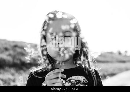 Süßes kleines Mädchen mit einer Dandelionenblume auf dem Land während eines sonnigen Frühlingstages - weibliches Kind, das Spaß hat und die Samen von Dandelion im Freien bläst - Stockfoto