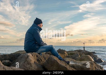 Mann, der allein auf Felsen sitzt und auf das Meer blickt. Stockfoto