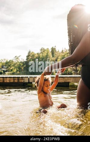Glücklicher Junge, der im Wasser genießt, während er im Urlaub die Hände der Mutter hält Stockfoto