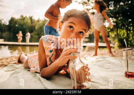 Mädchen schaut weg, während sie während des Urlaubs Saft aus der Flasche trinkt, die auf der Picknickdecke liegt Stockfoto