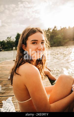 Seitenansicht einer lächelnden Frau, die während des Urlaubs auf dem Steg am See sitzt Stockfoto