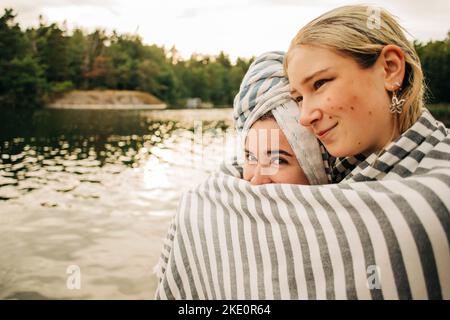 Frau umarmt weibliche Freundin in Handtuch eingewickelt am See während des Urlaubs Stockfoto