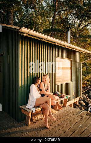 Weibliche Freunde sitzen auf der Bank in der Nähe der Hütte während des Sonnenuntergangs Stockfoto