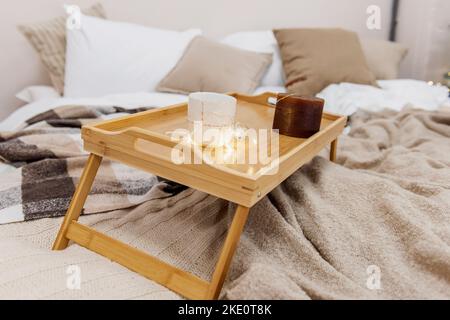 Neutrale Farben, monochromes Schlafzimmer. Ein Nachttisch aus Holz steht auf einem strukturierten Bettüberwurf mit kariertem Design. Auf dem Tisch stehen braune und milchige Kerzen mit garlan Stockfoto