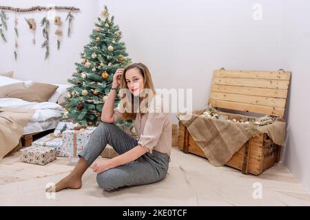 Stilvolle, moderne Millennial Frau sitzt in der Nähe Künstlicher Weihnachtsbaum, Holzkiste, Brust mit Glitzerkugeln. Festliche, umweltfreundliche Innenausstattung, mit festlichem Stockfoto