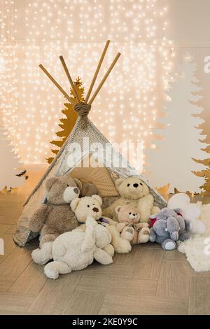 Kinderfotozone im Weihnachts-Fotostudio - Wigwam mit Teddybären auf dem Hintergrund von Silhouetten aus Tannenbäumen und Girlanden. Stockfoto