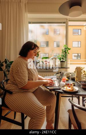 Junge Frau mit Laptop, während sie auf einem Stuhl am Tisch sitzt Stockfoto