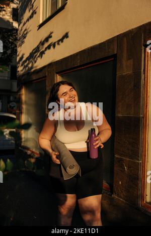 Glückliche junge Frau mit Übungsmatte und Wasserflasche im Hinterhof Stockfoto