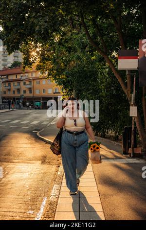 Die ganze Länge der Frau mit Einkaufstasche spricht auf dem Smartphone, während sie während des Sonnenuntergangs auf dem Fußweg läuft Stockfoto