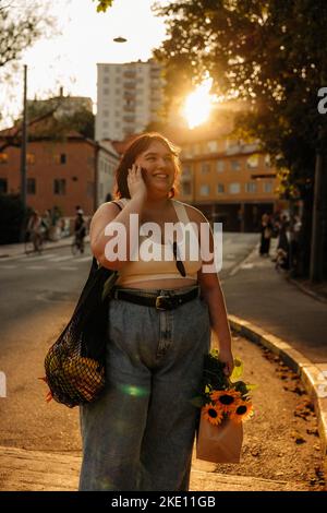 Glückliche, üppige Frau, die während des Sonnenuntergangs auf dem Smartphone spricht, während sie auf der Straße steht Stockfoto