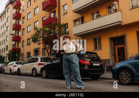 Porträt einer jungen selbstbewussten Frau, die mit der Hand auf der Hüfte gegen Autos steht Stockfoto