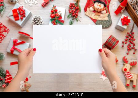 Frau hält ein leeres Papierblatt, um Grußkartentext hinzuzufügen. Weihnachtsszene mit vielen Geschenken und Dekorationen auf Holztisch Stockfoto
