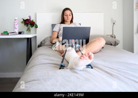 Junge Frau mit gebrochenem Bein auf dem Bett liegend, während sie einen Laptop benutzt Stockfoto