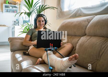 Junge Frau mit gebrochenem Bein auf dem Bett liegend, während sie einen Laptop benutzt Stockfoto