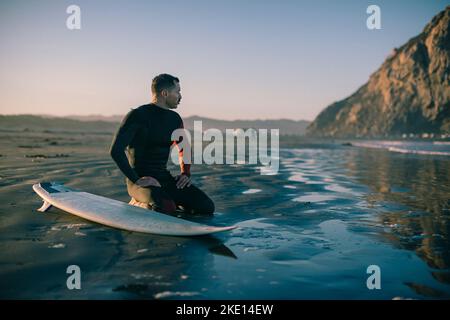 Ein Surfer kniet am Strand neben einem Surfbrett. Stockfoto