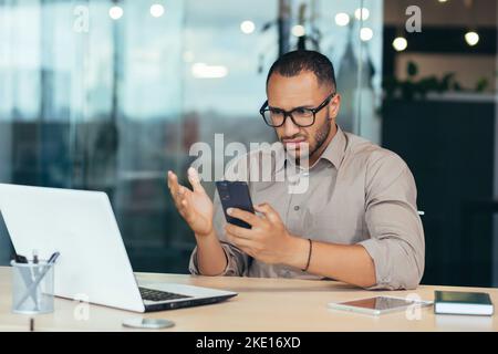 afroamerikanischer Mann erhielt online eine schlechte Nachrichtenmeldung vom Telefon, Geschäftsmann im Hemd, der mit einem Laptop mitten in einem modernen Bürogebäude arbeitete, Mann in einer Brille, der den Online-Brief las. Stockfoto