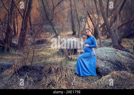 Ein schwanger Mädchen in einem blauen Kleid sitzt zwischen den Bäumen Stockfoto