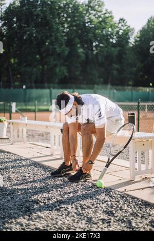 Mann, der Schuhspitze schnürt, während er auf der Bank auf dem Tennisplatz sitzt Stockfoto