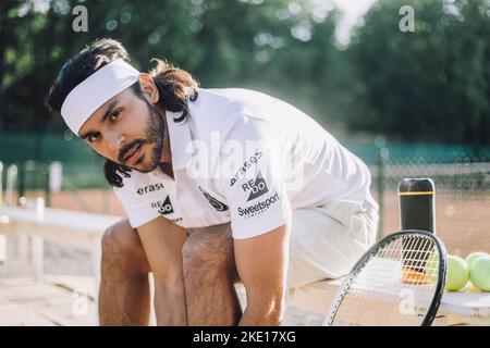 Selbstbewusster Mann mit Stirnband, während er auf der Bank auf dem Tennisplatz sitzt Stockfoto