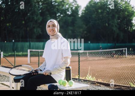 Porträt einer Frau mit Kopftuch, die einen Tennisschläger hält, während sie auf der Bank auf dem Sportplatz sitzt Stockfoto