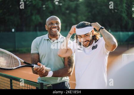 Glücklicher Mann mit Tennisschläger und einem Freund, der das Stirnband auf dem Sportplatz justiert Stockfoto