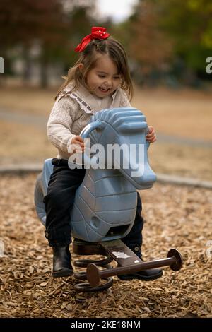 Entzückendes, 3 Jahre altes kleines Mädchen, das im Herbst in einem Stadtpark in British Columbia, Kanada, auf einem Spielplatz reitet und fröhlich lächelt Stockfoto