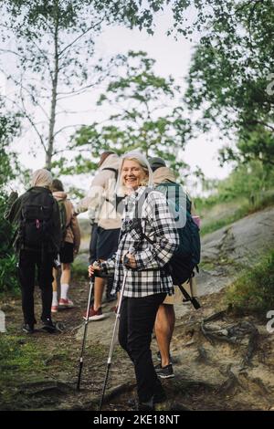 Seitenansicht einer lächelnden älteren Frau, die beim Wandern mit Freunden im Wald die Stange hält Stockfoto