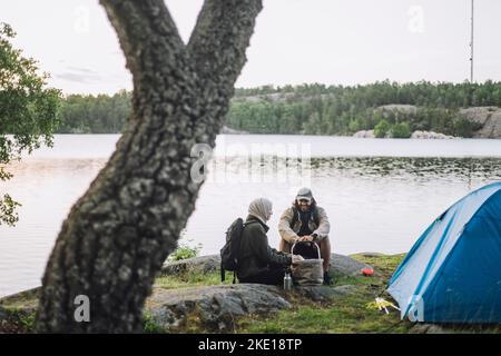 Freunde unterhalten sich miteinander, während sie am Seeufer auf Felsen am Zelt sitzen Stockfoto
