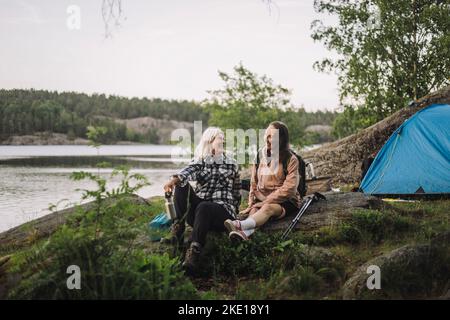 Lächelnde ältere Frauen sprechen miteinander, während sie während des Campens auf Felsen sitzen Stockfoto