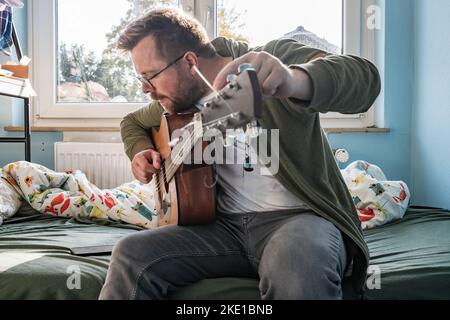 Akustische Gitarre Tuning. Ein Mann repariert ein Saiteninstrument, während er auf ein Smartphone schaut. Stockfoto