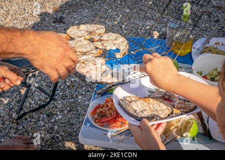 Eine Frau mit einer Gabel, Schwertfischscheiben auf einem weißen Teller und einem grill. Selektiver Fokus auf einem Plastiktisch mit Zitronenfrucht und Garnelenköpfen Stockfoto