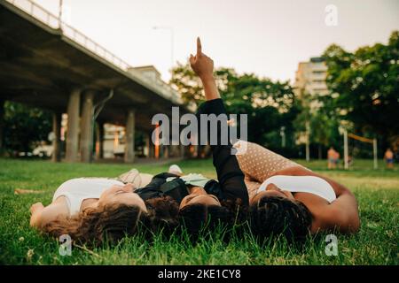 Teenager-Mädchen mit erhobener Hand zeigt, während sie inmitten von Freunden auf dem Gras im Park liegt Stockfoto