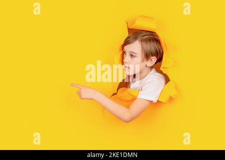 Junge in rotem Papierloch, der mit dem Finger zur Seite zeigt. Lächle Junge, der durch ein Loch in der Papierwand schaut. Überraschte das Kind, das Loch in Papier machte. Blonde Jungen sieht aus Stockfoto