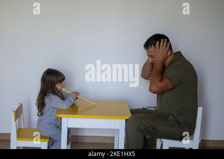 Kleines Mädchen spielt Flöte und Vater stopft seine Ohren. Musikunterricht eines kleinen Mädchens, das spielen lernt. Stockfoto