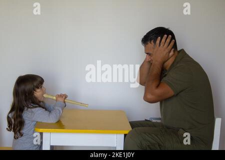 Kleines Mädchen spielt Flöte und Vater stopft seine Ohren. Musikunterricht eines kleinen Mädchens, das spielen lernt. Stockfoto