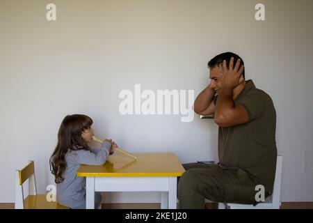 Kleines Mädchen spielt Flöte und Vater stopft seine Ohren. Musikunterricht eines kleinen Mädchens, das spielen lernt. Stockfoto