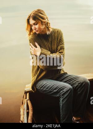 Eine traurige, schöne Frau in einem Wollpullover sitzt am Rand eines Holzbootes, das auf der Wasseroberfläche des Ozeans schwimmt. Wasser und Ruhe. Stockfoto