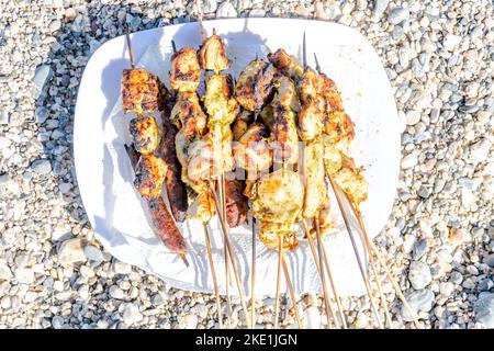 Aus nächster Nähe sehen Sie einen weißen Teller voller marinierter Hühnerspieße und gegrillter Merguez. Natürliches Sonnenlicht mit Schatten und Kieselsteinen am Strand Stockfoto