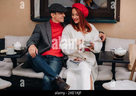 Ein glückliches, stilvolles Paar trinkt Kaffee und lächelt, während es in einem Café auf der Straße sitzt Stockfoto