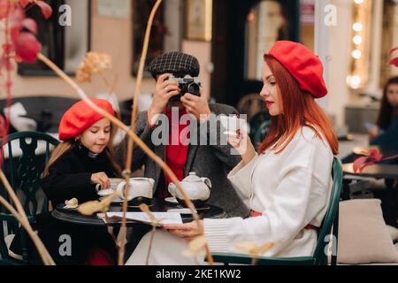 Eine stilvolle dreiköpfige Familie sitzt draußen an einem Tisch in einem Café und trinkt Kaffee. Papa fotografiert Mama in der Herbststadt Stockfoto